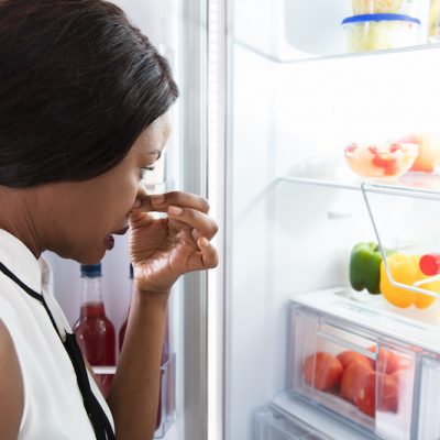 Close-up Of An African Young Woman Holding Her Nose Near Foul Food In Refrigerator